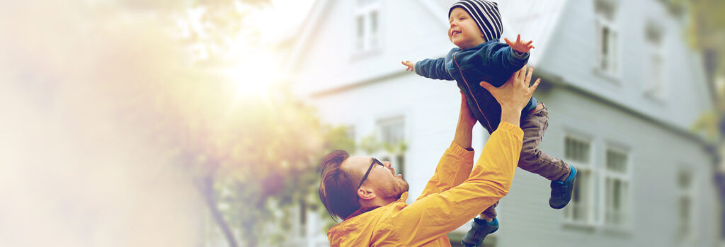 Father lifting his laughing infant son in the air in front of a two-storey home.